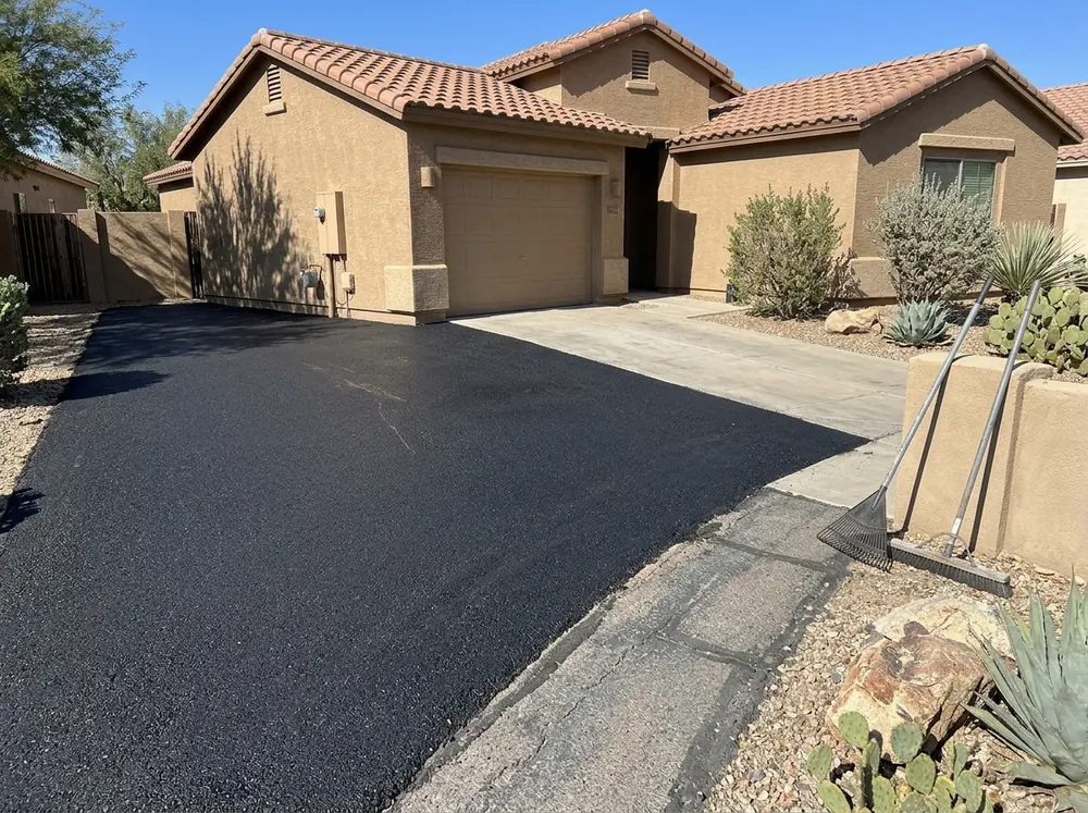 Fresh black asphalt overlay on a residential driveway in front of a stucco home, the edge of the old gray surface still visible at the side