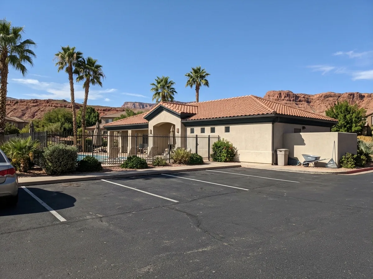 Small asphalt parking lot beside a stucco clubhouse with a pool fence, palm trees and red rock hills behind