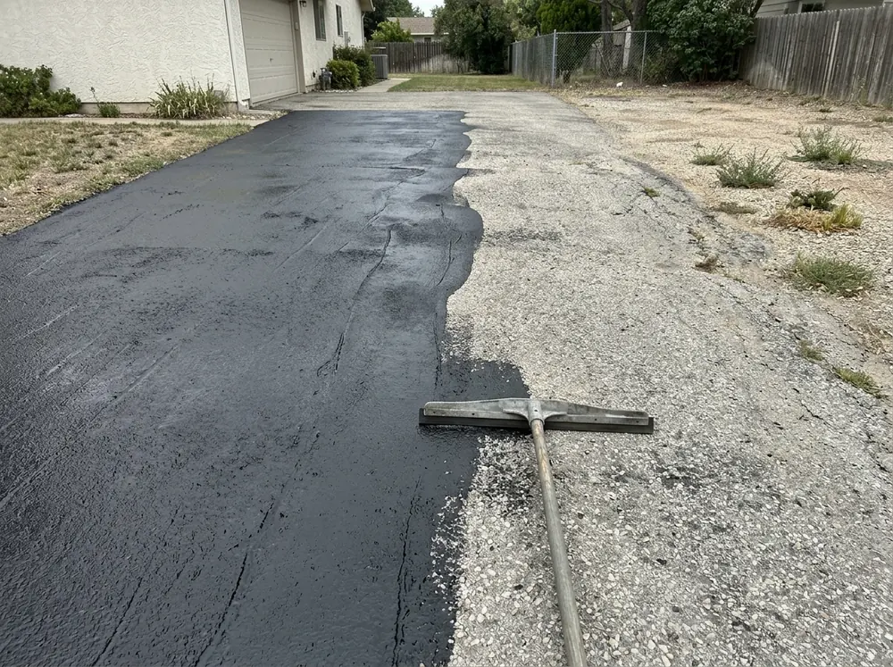 Worn, porous gray asphalt driveway half covered with a thick black fill-and-seal coating
